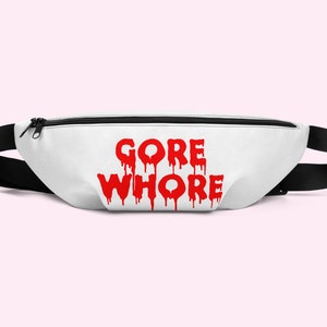 Puede incluir: Riñonera blanca con el texto rojo "GORE WHORE" impreso en la parte delantera. La bolsa tiene una correa negra ajustable y un cierre de cremallera negro. La riñonera está diseñada para llevarse alrededor de la cintura.