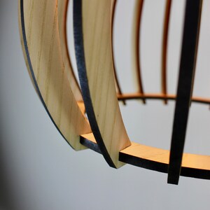 May include: Close-up of a wooden pendant light fixture with a natural wood finish and black accents. The light fixture is made of thin strips of wood that are woven together to create a circular shape.