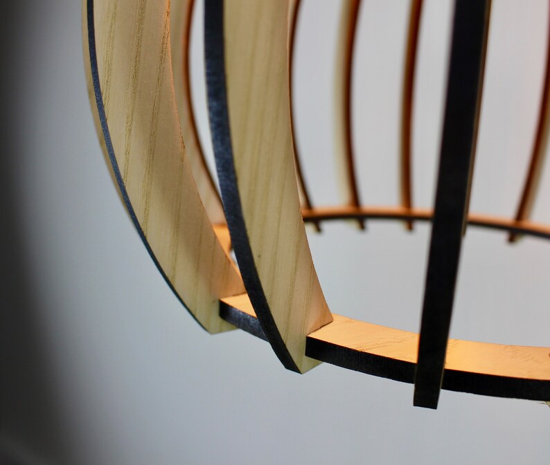 May include: Close-up of a wooden pendant light fixture with a natural wood finish and black accents. The light fixture is made of thin strips of wood that are woven together to create a circular shape.