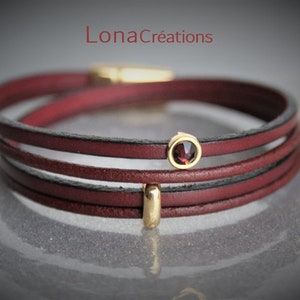 Pode incluir: Um bracelete de couro bordô com três fios e um detalhe dourado. Uma pequena gema redonda de cor vermelho escuro está engastada em uma moldura dourada.