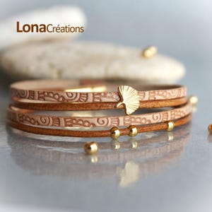 Könnte beinhalten: Ein dreifach gewickeltes Lederarmband mit einem goldenen Ginkgoblatt-Anhänger und goldenen Perlen. Das Leder ist hellbraun und hat ein gemustertes Design.