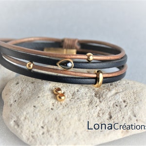 May include: A multi-layered leather bracelet with black and brown leather straps, gold beads, and a small black gemstone. The bracelet is on a light-colored stone surface. LonaCréations is written on the surface.