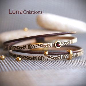 May include: A multi-layered bracelet with brown leather bands and a white band with a gold embossed pattern. A small red gemstone is set in a gold bezel on the white band.
