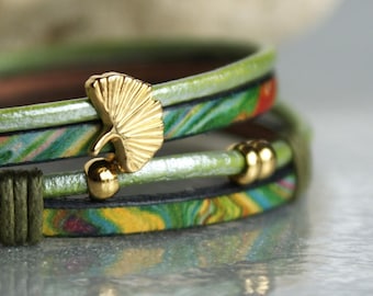 May include: A multi-layered bracelet with a green, blue, and yellow patterned leather strap. The bracelet has a gold ginkgo leaf charm and gold beads.