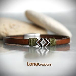 Peut inclure: Un bracelet en cuir marron avec un charme géométrique argenté et des accents de cordon vert. Le charme présente un motif de croix.