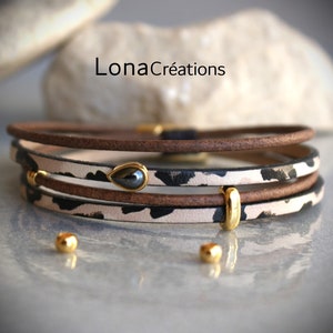 Könnte beinhalten: Ein mehrlagiges Lederarmband mit einem schwarz-weißen Tiermuster. Das Armband ist mit einer goldenen Perle und einem tropfenförmigen schwarzen Edelstein verziert.