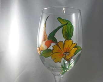 May include: A clear wine glass with a hand-painted design featuring a yellow flower, green leaves, and a small orange and white bird. The stem and base are clear glass.