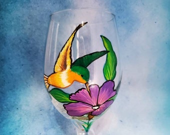 May include: Hand-painted wine glass with a hummingbird in flight, sipping nectar from a purple flower with green leaves.