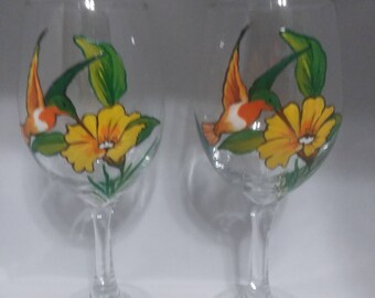 May include: Two clear glass wine glasses with hand-painted hummingbird and flower designs. The flowers are yellow with green leaves and the hummingbirds are orange, white, and green.