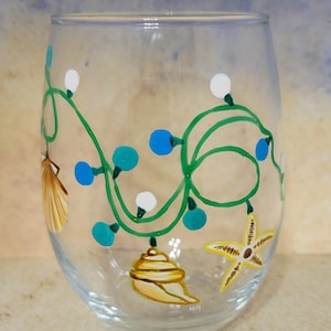 May include: A clear, stemless wine glass hand-painted with a green vine design featuring white and blue accents, and seashell and starfish illustrations. The text "Hand painted by Marescalt" is at the bottom.