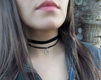 May include: A black velvet choker necklace with a silver crescent moon pendant. The necklace is worn over a denim jacket and a white top. The choker is a stylish accessory.
