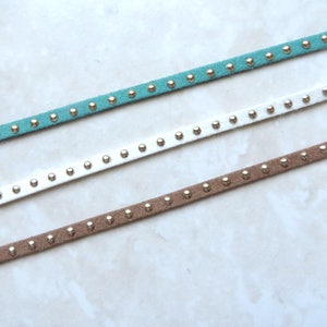 Puede incluir: Tres cordones de ante de diferentes colores con tachuelas plateadas. Los cordones son azul verdoso, blanco y marr&oacute;n.