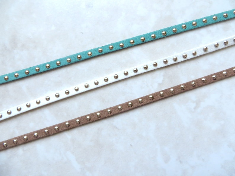 Puede incluir: Tres cordones de ante de diferentes colores con tachuelas plateadas. Los cordones son azul verdoso, blanco y marr&oacute;n.