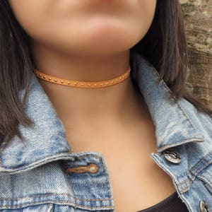 Puede incluir: Un collar gargantilla de cuero marr&oacute;n claro con tachuelas plateadas. La gargantilla la lleva una persona con el pelo largo y oscuro y una chaqueta vaquera.