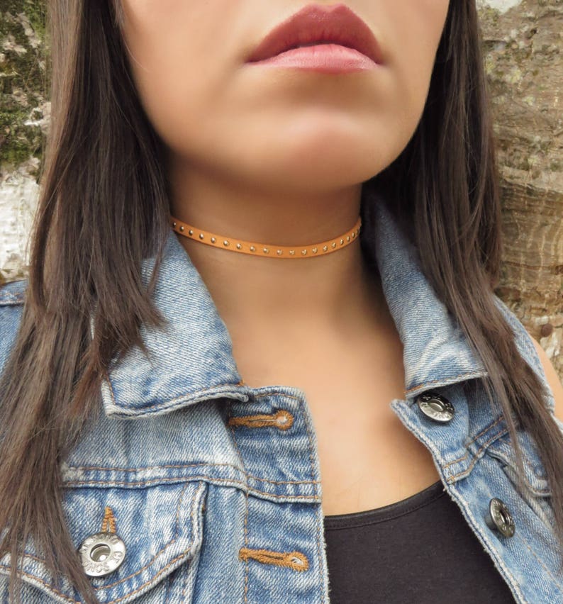 Puede incluir: Un collar gargantilla de cuero marr&oacute;n con tachuelas plateadas. El collar es delgado y tiene un dise&ntilde;o simple.