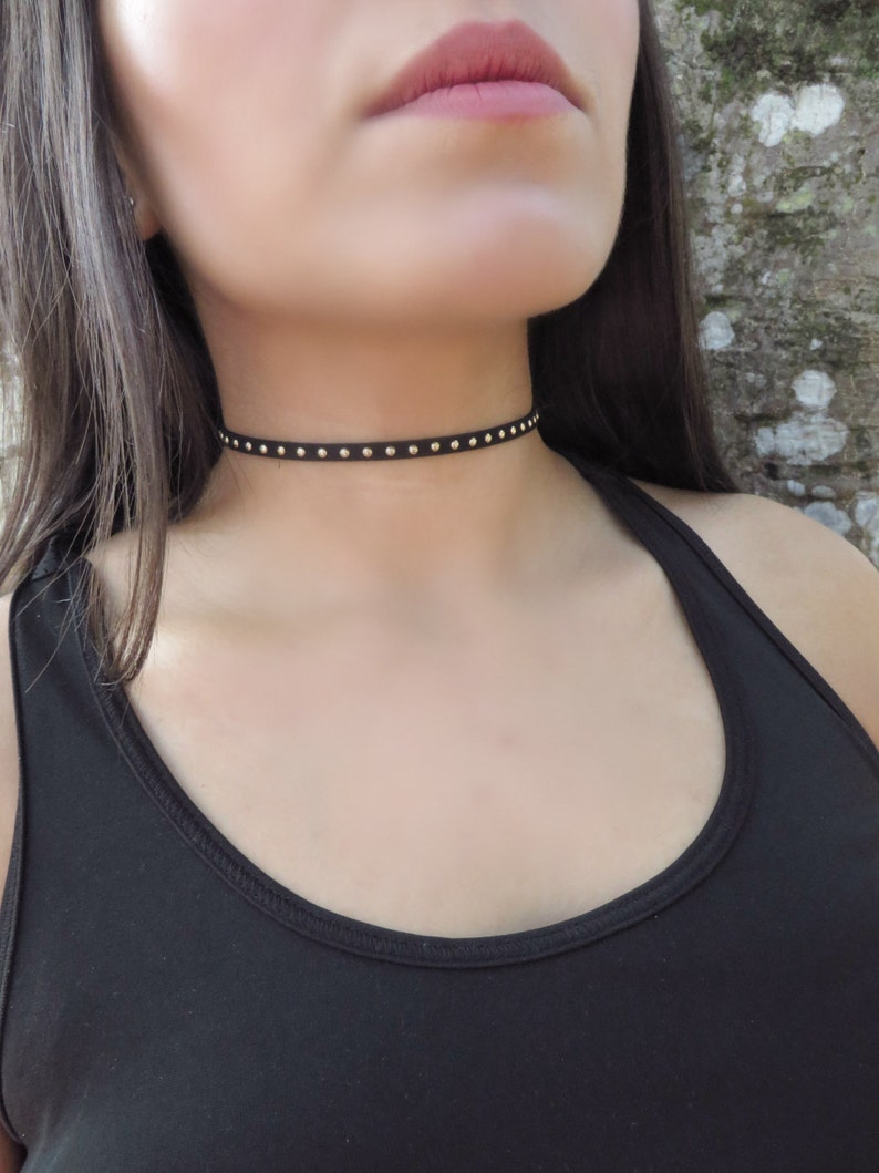 Puede incluir: Un collar gargantilla negro con tachuelas doradas. El collar es fino y delicado, y las tachuelas est&aacute;n espaciadas uniformemente.