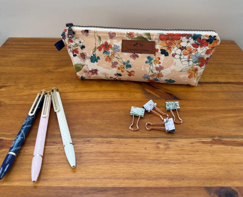 May include: A floral print pencil case with a zipper closure, pens, and binder clips on a wooden surface. The pencil case features a beige background with colorful flowers. Three pens are visible, along with several binder clips.
