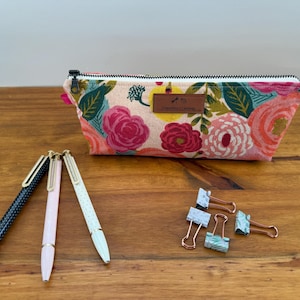 May include: A floral print pencil case with a zipper closure, featuring pink, red, and yellow flowers. Three pens and several binder clips are arranged on a wooden surface. The pens are black, pink, and white.