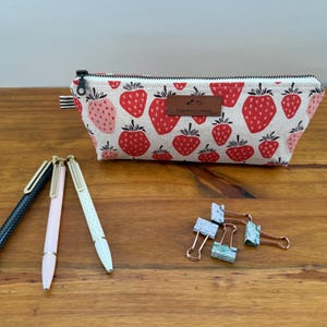 May include: A canvas pencil case with a strawberry pattern, a black zipper, and a small brown label. Three pens and several binder clips are arranged on a wooden surface.
