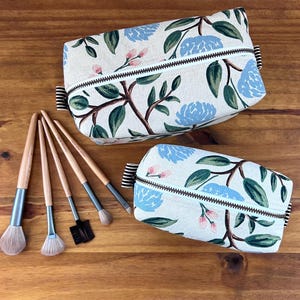 May include: Two floral print cosmetic bags with a light blue and pink flower design, and a set of makeup brushes with wooden handles. The bags have a zipper closure and are rectangular in shape.