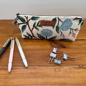 May include: A floral print pencil case with a zipper closure, a leather tag, and three pens. The pens are black, pink, and white. Also, there are four binder clips on a wooden surface.
