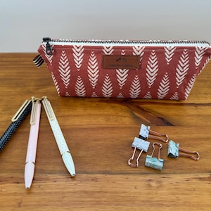 May include: A red pencil case with a white leaf pattern, a black zipper, and a small leather tag. Three pens and three binder clips are arranged on a wooden surface. The pens are black, pink, and white with gold accents.
