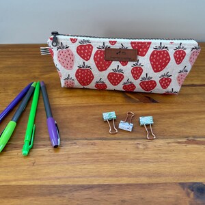 May include: A rectangular pencil case with a strawberry pattern, a black zipper, and a brown leather tag. Three pens and three paper clips are on a wooden surface.