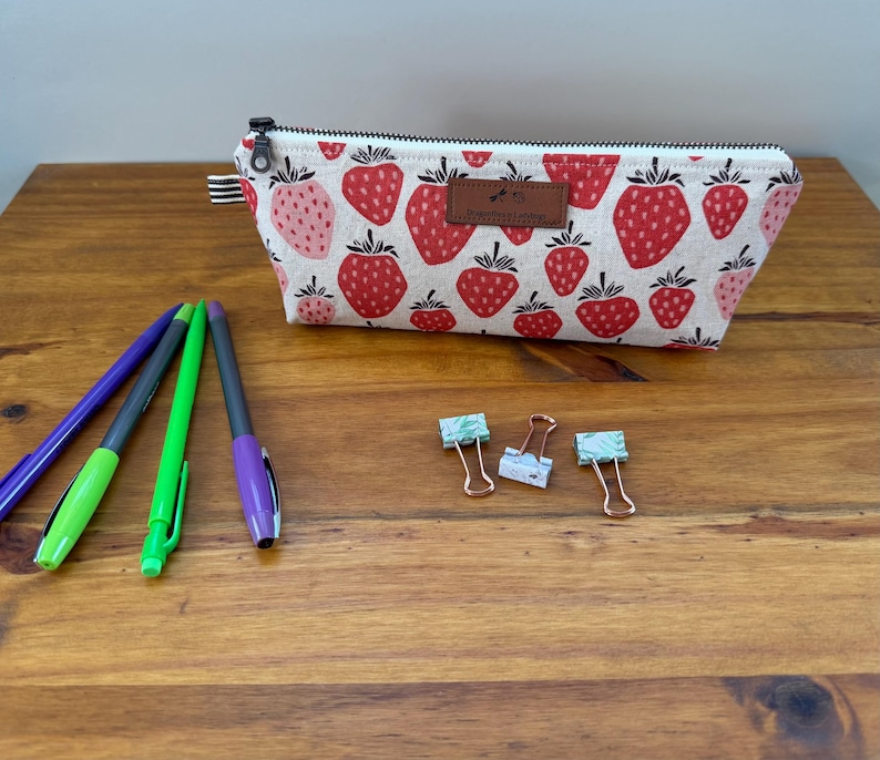 May include: A rectangular pencil case with a strawberry pattern, a black zipper, and a brown leather tag. Three pens and three paper clips are on a wooden surface.