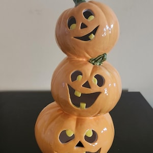 May include: A vertical stack of three orange ceramic pumpkins with carved jack-o'-lantern faces. Each pumpkin features cutouts for eyes, a triangular nose, and a wide mouth with visible teeth, finished with a glossy glaze and green stems.