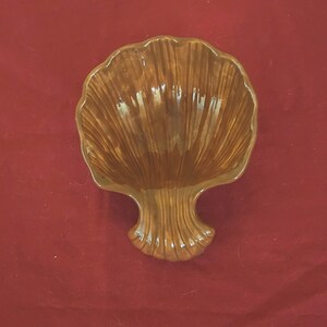 May include: A brown, shell-shaped ceramic dish with a pedestal base. The dish has a glossy finish and is set against a red background. The shell design features vertical ridges.