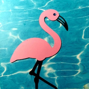 May include: Pink paper flamingo cutout with black legs and a blue jewel eye, standing on a blue background with white ripples.