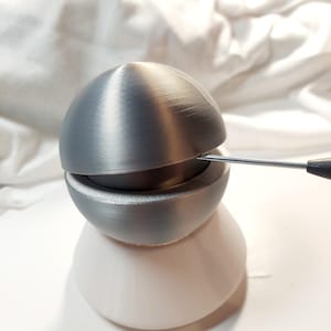 May include: A silver-colored, two-piece, 3D printed egg-shaped container with a small opening on the top. A thin metal tool is being used to pry open the top half of the egg.