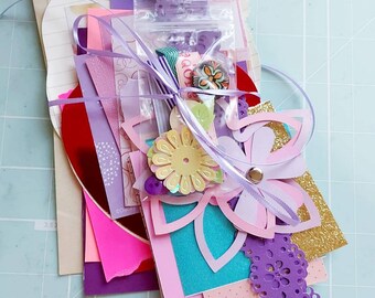 May include: A collection of purple, pink, and teal paper crafting supplies, including patterned paper, lace, flowers, and a small bag of embellishments. The supplies are tied together with a purple ribbon and are arranged on a light blue surface.