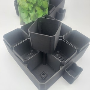 May include: Black 3D-printed planter with multiple square compartments arranged in a geometric pattern. A small square pot is detached. A packet of cilantro is in the background. The planter is designed for small plants or herbs.