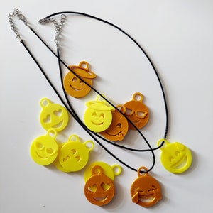 May include: A black necklace with multiple yellow and orange emoji charms. The charms include a smiling face with a halo, a winking face, a heart-eyed face, a face with sunglasses, a face with tears of joy, and a face with three hearts.