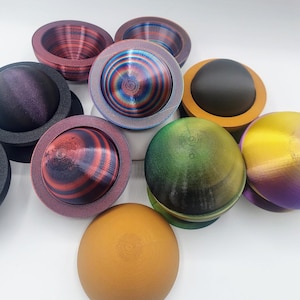 May include: A collection of colorful, spherical objects with a planet-like appearance. The spheres feature various color schemes, including red, blue, green, and gold, with textured surfaces and ring-like bases. The objects are arranged on a white surface.