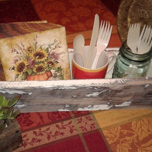 May include: A rustic wooden box with a distressed white finish, containing a decorative wooden block with a sunflower and pumpkin design, a red and white mug with plastic utensils, and a glass jar with plastic forks.