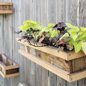 Puede incluir: Dos macetas de madera montadas en una valla de madera gris. La maceta superior está llena de plantas verdes y moradas. La maceta inferior está vacía.