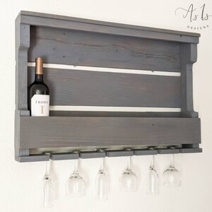 May include: Gray wooden wall mounted wine rack with six glass holders and a shelf for storing bottles. The rack has a white painted stripe across the top and bottom shelves. A bottle of wine is on the top shelf with the label "IRONY" visible.