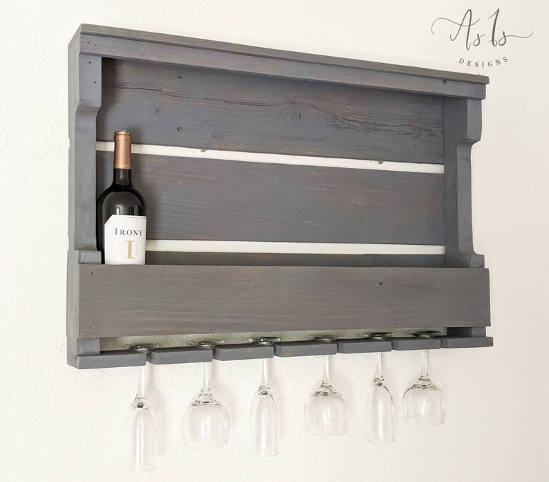 May include: Gray wooden wall mounted wine rack with six glass holders and a shelf for storing bottles. The rack has a white painted stripe across the top and bottom shelves. A bottle of wine is on the top shelf with the label "IRONY" visible.