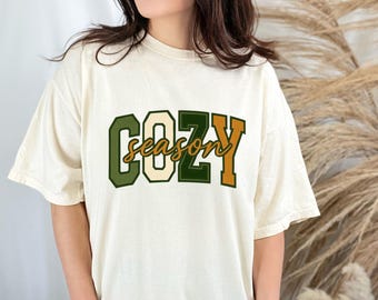 May include: Cream-colored t-shirt with the words "COZY SEASON" in a collegiate-style font. The text is in shades of green, cream, and orange. The shirt has a relaxed fit and short sleeves.