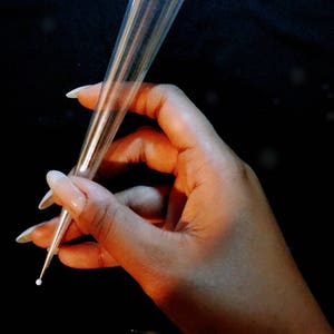 May include: A clear, cone-shaped applicator with a metal tip, held against a dark background. The applicator is transparent, and the tip has a small white dot. The image focuses on the applicator.