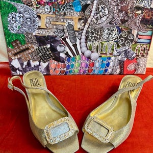 May include: A pair of gold-colored slingback sandals with rectangular, jeweled buckles. The sandals have a metallic sheen and are set against a red background. The brand name "Pelle" is visible on the insole.