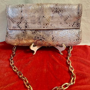 Op de afbeelding: Een zilveren metallic clutch met een slangenhuidpatroon en een gouden kettingband.