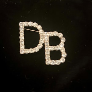 Könnte beinhalten: Eine silberfarbene Brosche mit den Buchstaben "DB" in Strasssteinen.