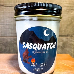 Könnte beinhalten: Ein weißes Kerzengefäß mit einem blauen und weißen Etikett, auf dem ein Comic-Sasquatch zu sehen ist, der Marshmallows über einem Lagerfeuer brät. Das Etikett enthält auch den Text "Sasquatch" und "Fir, Evergreens, Cedar, Moss". Am unteren Rand des Etiketts steht "Luna Love Candles".