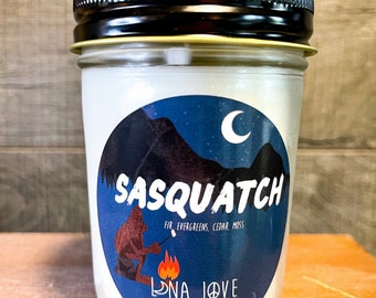 May include: A white candle jar with a blue and white label featuring a cartoon Sasquatch roasting marshmallows over a campfire. The label also includes the text "Sasquatch" and "Fir, Evergreens, Cedar, Moss". The bottom of the label reads "Luna Love Candles".