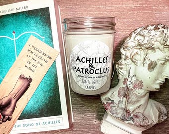 May include: A glass jar candle with a marble-patterned label reading Achilles & Patroclus, positioned next to a book titled The Song of Achilles, a wooden bookmark with a hand illustration and text, and a weathered white bust of a male figure.