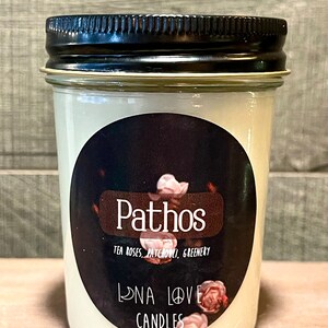 Könnte beinhalten: Eine weiße Kerze in einem Glasgefäß mit schwarzem Deckel. Das Etikett lautet "Pathos" und "Tea Roses, Patchouli, Greenery" sowie "Lona Love Candles".