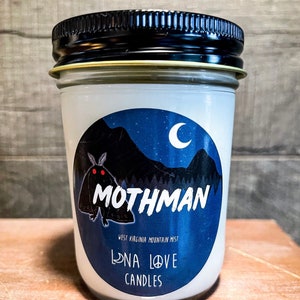 Könnte beinhalten: Ein weißes Kerzengefäß mit einem blauen und schwarzen Etikett, das eine Mothman-Illustration, einen zunehmenden Mond und den Text "Mothman", "West Virginia Mountain Mist", "Luna Love Candles" zeigt.